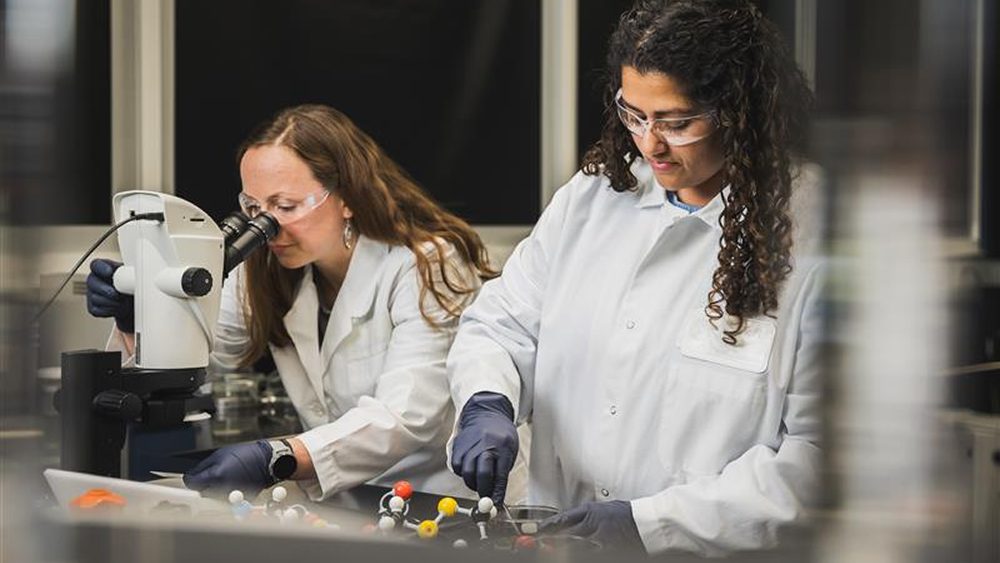 Two scientists in lab coats conducting research, one looking through a microscope and the other working with laboratory equipment.