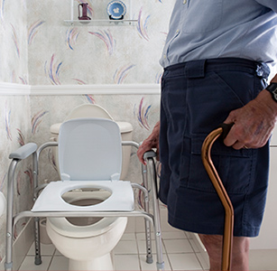 Man holding a cane, standing next to a toilet with a raised toilet seat.