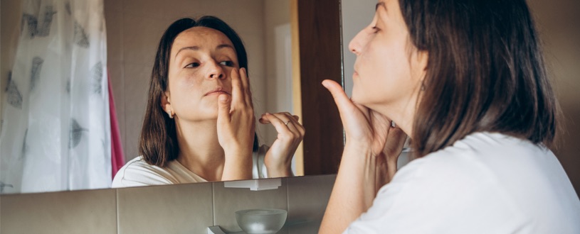 A woman uses her bathroom mirror to apply a facial serum under her eyes.