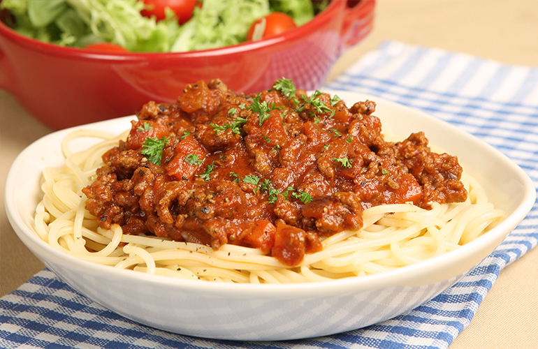 espaguete a bolonhesa em um prato branco com mesa e um bowl vermelho com salada ao lado