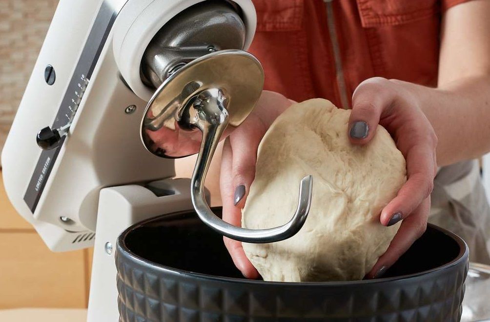 Batedeira Artisan Branca com Bowl edição limitada preto com detalhes em relevo. Uma mão delicada com esmalte roxo, aparamente feminina, coloca uma massa lisa na tigela da batedeira.