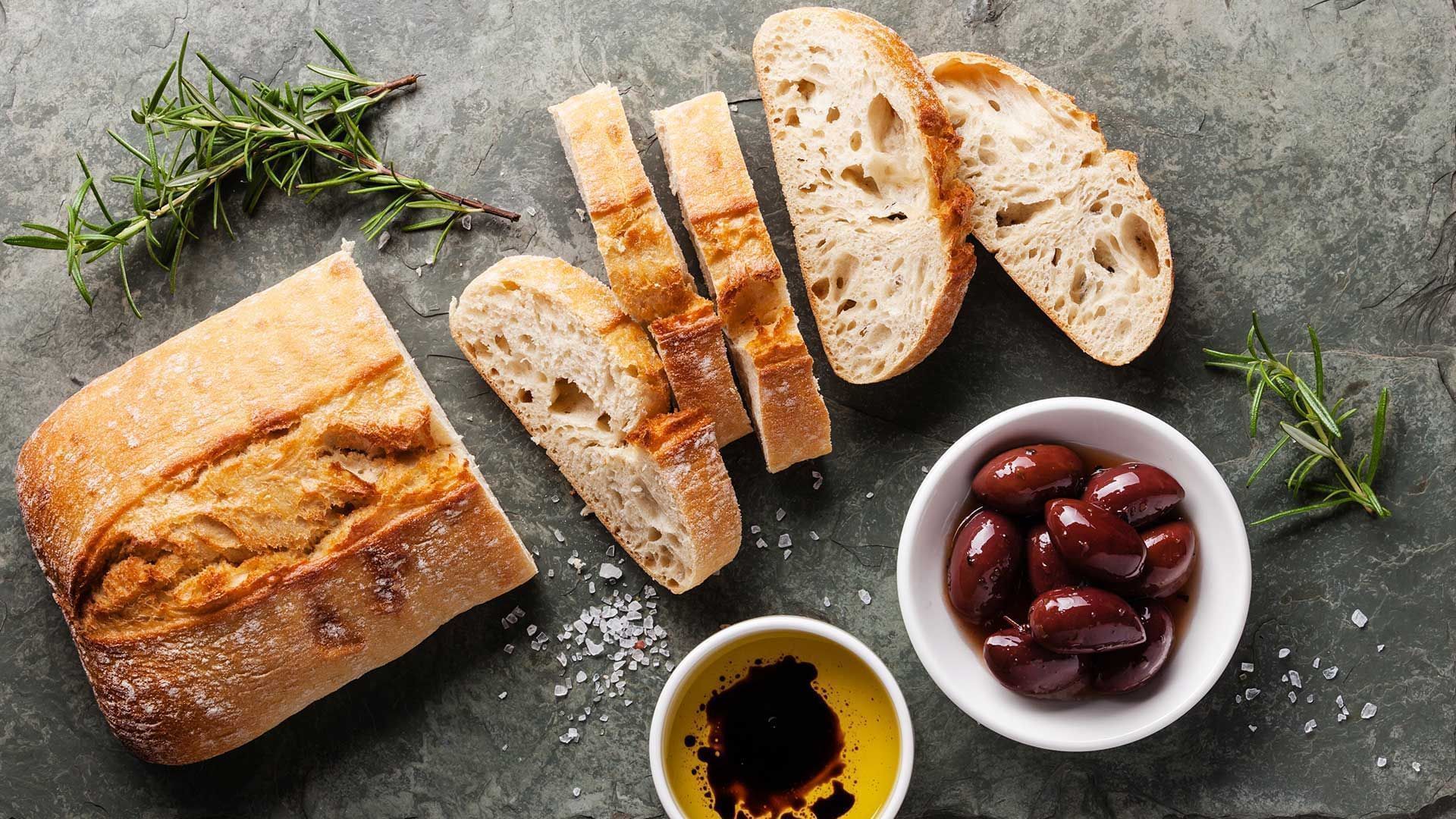 Em uma bancada de cor grafite vemos um pão cortado pela metade, em que uma das metades está cortado em fatias. No canto superior da tela vemos pequenos ramos de alecrim. No canto inferior da tela vemos uma tigela de azeite temperado, uma tigela de azeitonas pretas e também outro ramo de alecrim. Há farinha polvilhada na mesa próximo ao pão.