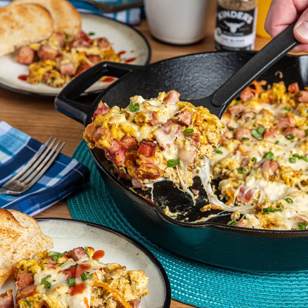 Hand taking scoop out of cast iron skillet filled with eggs ham and melted cheese