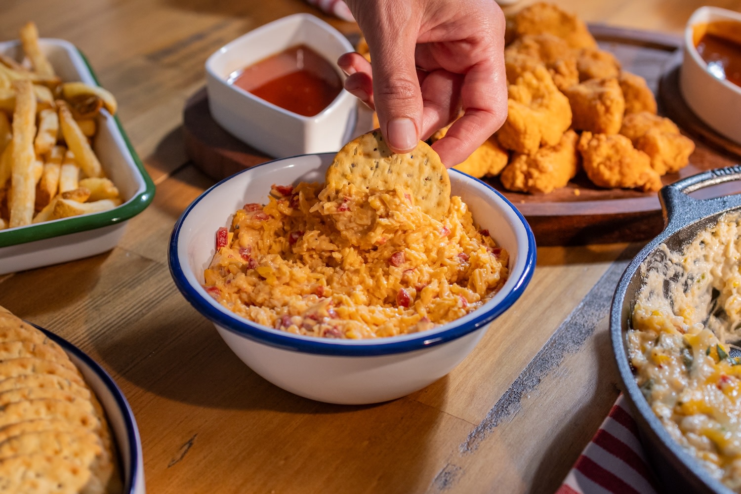 Pimento cheese dip in a white bowl featuring a hand dipping a cracker into the spread