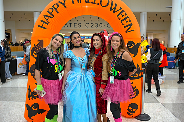 2025 Halloween at MCO photo of four women dressed in costume standing in front of a Happy Halloween blow up arch.