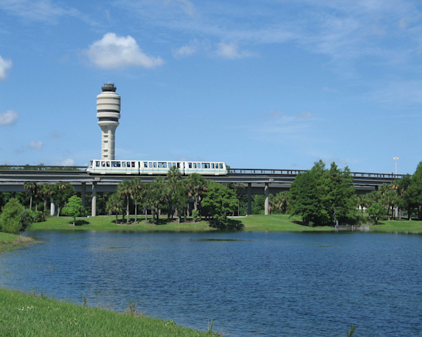 Outside photo of a lake in front of a Terminal Link, which is moving along the track, with the MCO Tower right behind it.