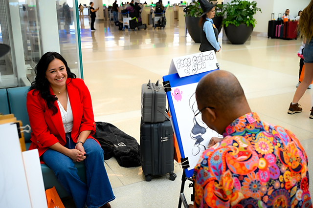 2025 Halloween at MCO photo of a lady wearing a red jacket sitting down on a couch getting a caricature drawing of herself done.