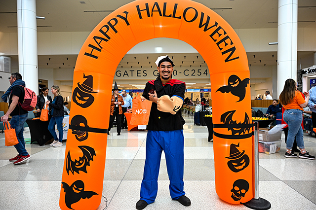 2025 Halloween at MCO photo of a man dressed up in a Popeye the Sailor costume standing under a orange "Happy Halloween"  arch.