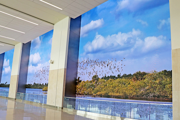Designed by: Gentilhomme Inc. & Sardi Designs
Details: Unique to Orlando International Airport, this immersive digital storytelling feature explores the knowns and unknowns of Central Florida, and captures the transformative development of the region. Windows on Orlando is composed of three identical LED displays, each 30 feet wide and 33 feet tall. They offer panoramic views into original live action scenes with a pixel pitch of 4.166 mm LED and a resolution of 19K. Experience a rocket launch filmed real-time from the Kennedy Space Center, an afternoon ride under open skies to round up cattle with cowhands at the Deseret Ranch, and a larger-than-life tribute to the art of the Florida Highwaymen and city of Orlando sculptural icons. The experience offers unexpected moments of magic to restore the joy and wonder of air travel.
Installation: Windows on Orlando
Location: Gates C230-C238