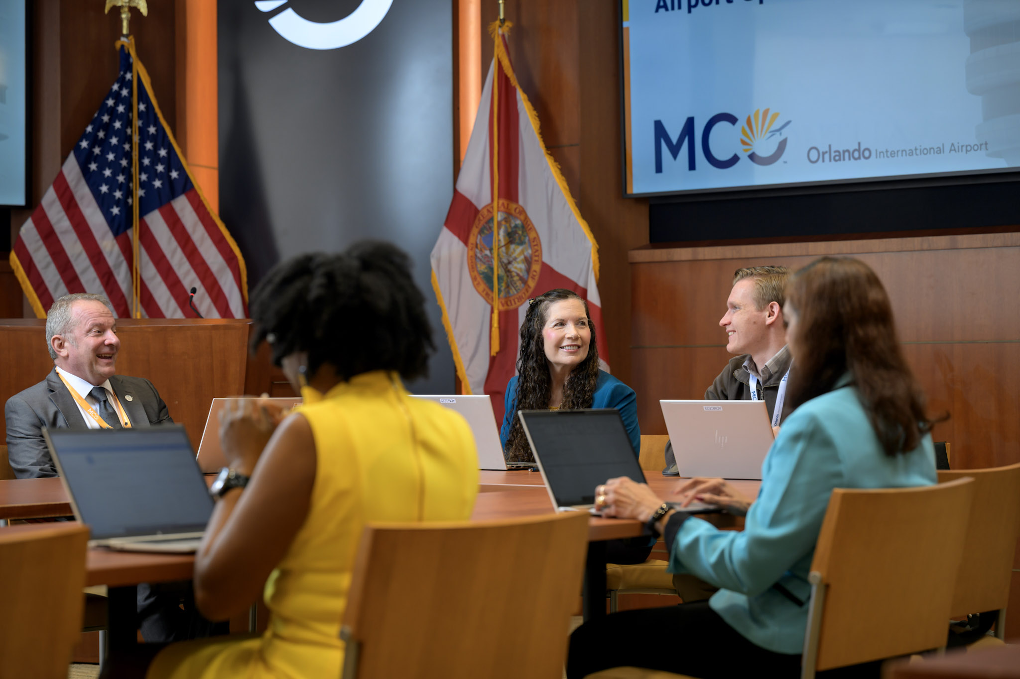 A group of people are in a board room gathered at table, talking and looking at their laptops.