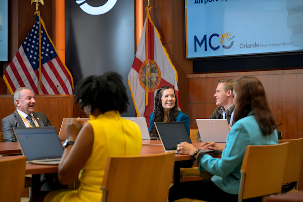 A group of people are in a board room gathered at table, talking and looking at their laptops.