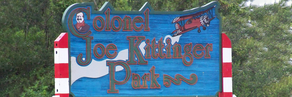 A wooden sign painted in red, white, and blue marks Colonel Joe Kittinger Park, an aviation-themed park dedicated in 1992. Located in the southwest corner of the airport off Crystal Lake Drive, the park honors hometown hero and aviation legend Colonel Joe Kittinger.