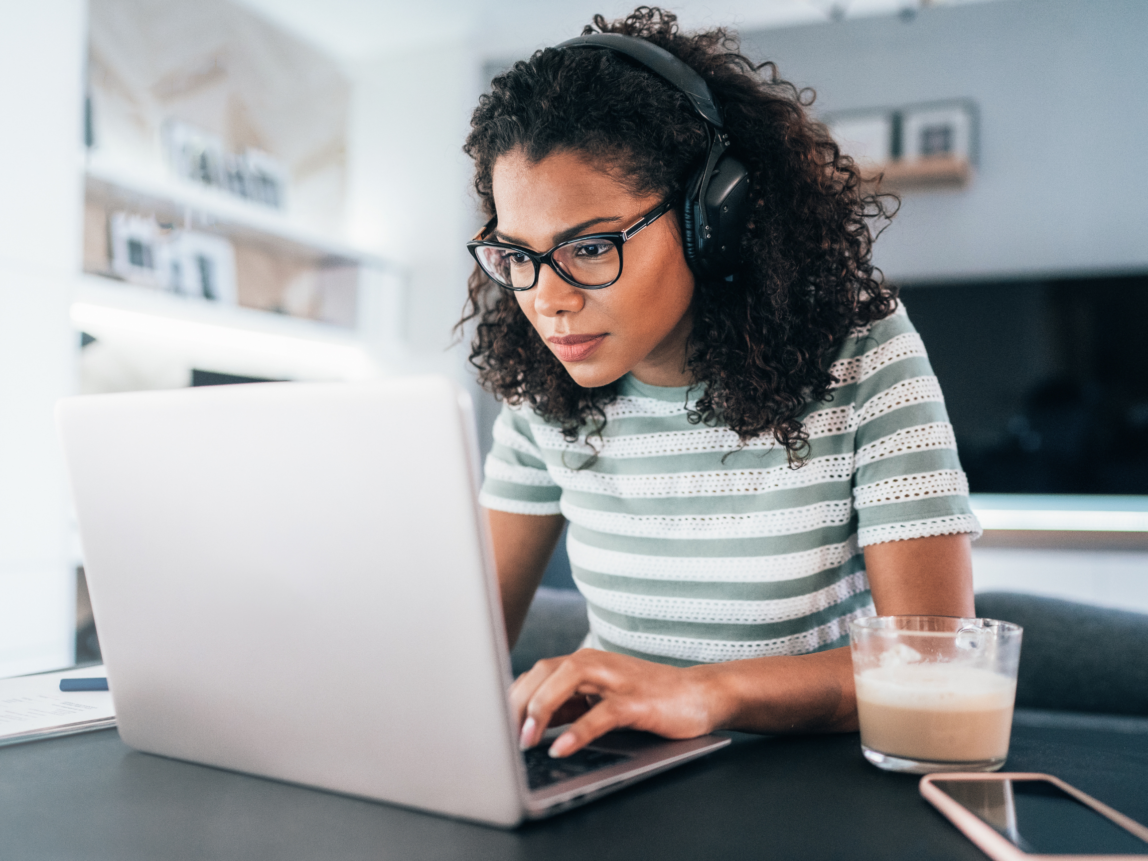 A young woman wearing glasses is at a desk wearing headphones looking at her silver laptop.