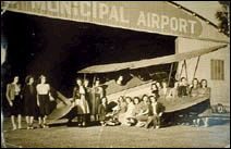 An old 1935 black and white photo of Amelia Earhart standing in front of a bi-plane parked in front of an Orlando Municipal Airport hangar.