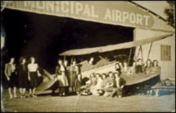 An old 1935 black and white photo of Amelia Earhart standing in front of a bi-plane parked in front of an Orlando Municipal Airport hangar.