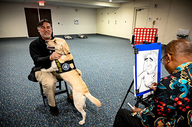 2025 Federal Airport Employee Appreciation Day photo of federal employee sitting down with a Paw Pilots dog sitting in his lap, while getting a caricature done.