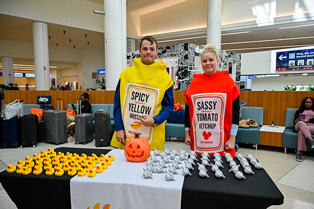 2025 Halloween at MCO photo of a couple dressed up as ketchup and mustard bottle standing behind a table of rubber duckies.