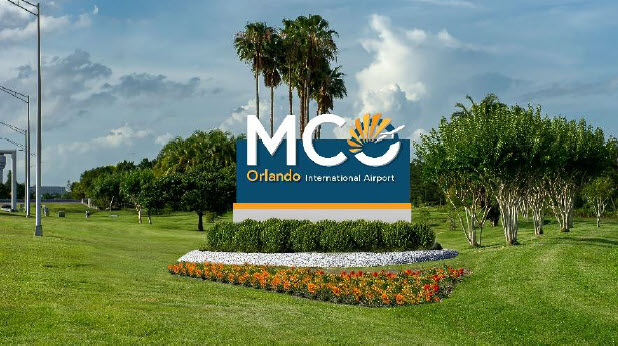 The MCO Orlando International Airport sign standing outside in front of palm tree with lush green grass all around it.