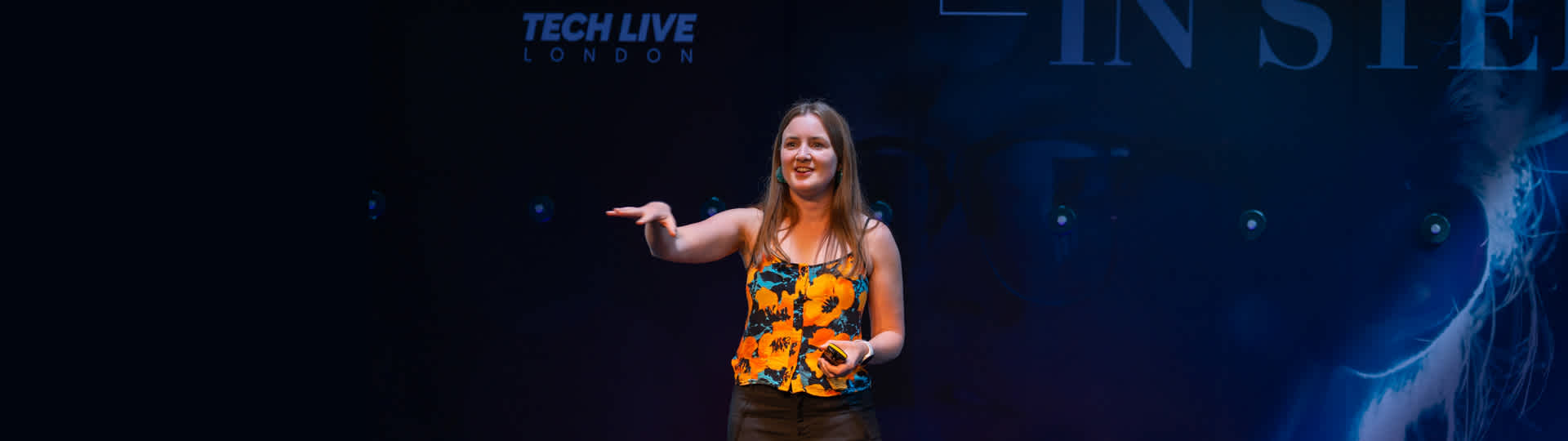 a woman in a poppy printed top standing on a stage. a sign in the background reads Tech Live London