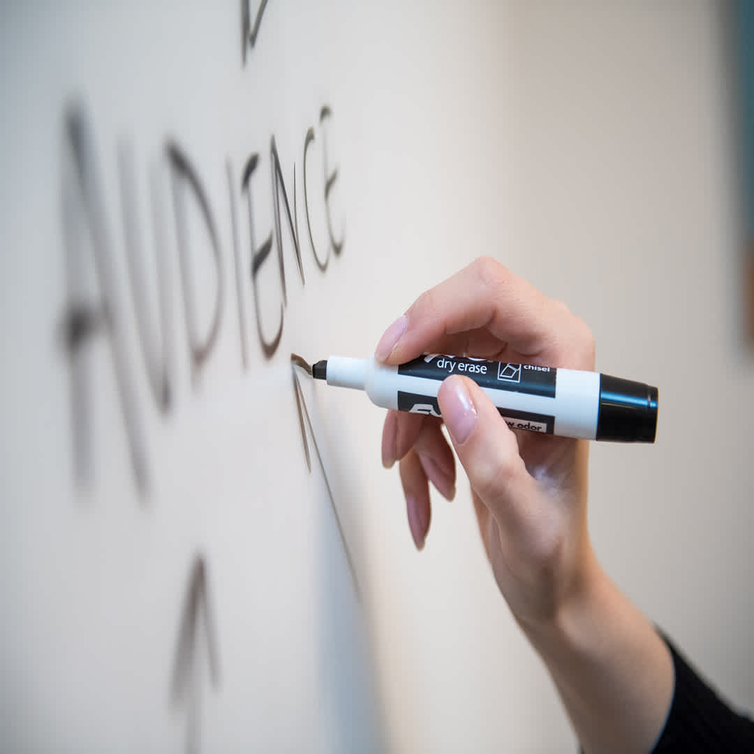 a hand holding a black coloured marker writing the word audience on a whiteboard