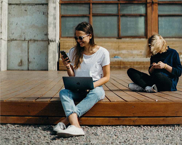 woman wearing sunglasses using smartphone and laptop sitting next to a person using a smartphone