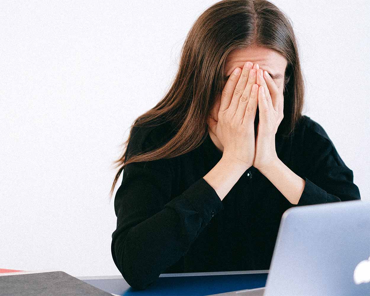 woman in black top in front of a laptop with her hands covering her face
