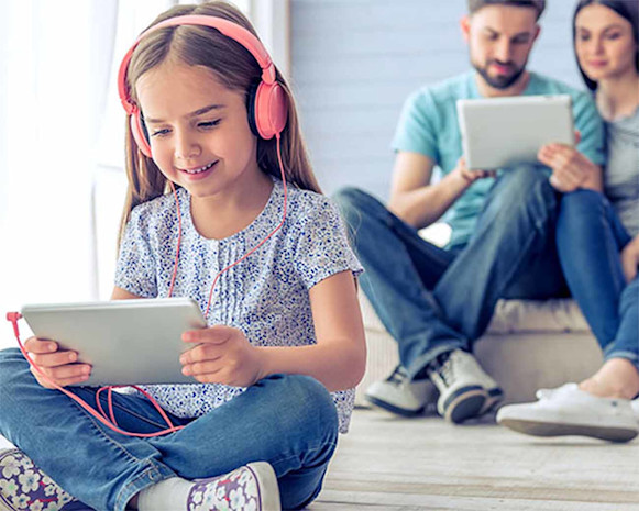 child wearing pink headphones holding tablet, with a man and a woman looking at a tablet in the background