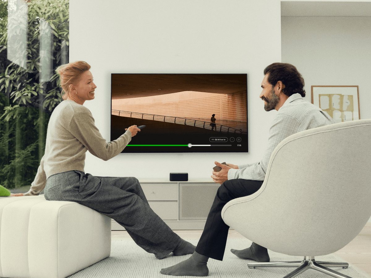 En mand og en kvinde kigger på en hinanden mens et tv kører i baggrunden. 