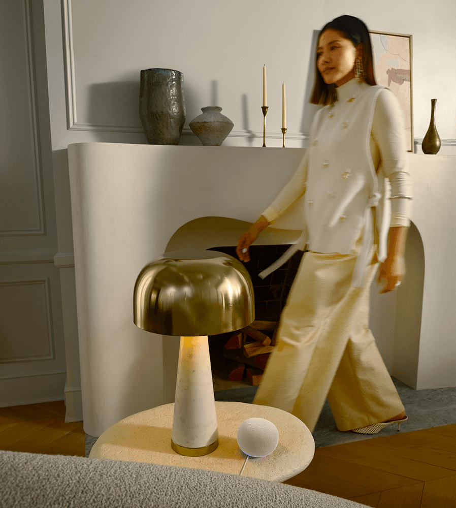 En hvid Google Home Speaker står på et lille bord ved siden af en lampe. I baggrunden går en kvinde forbi.