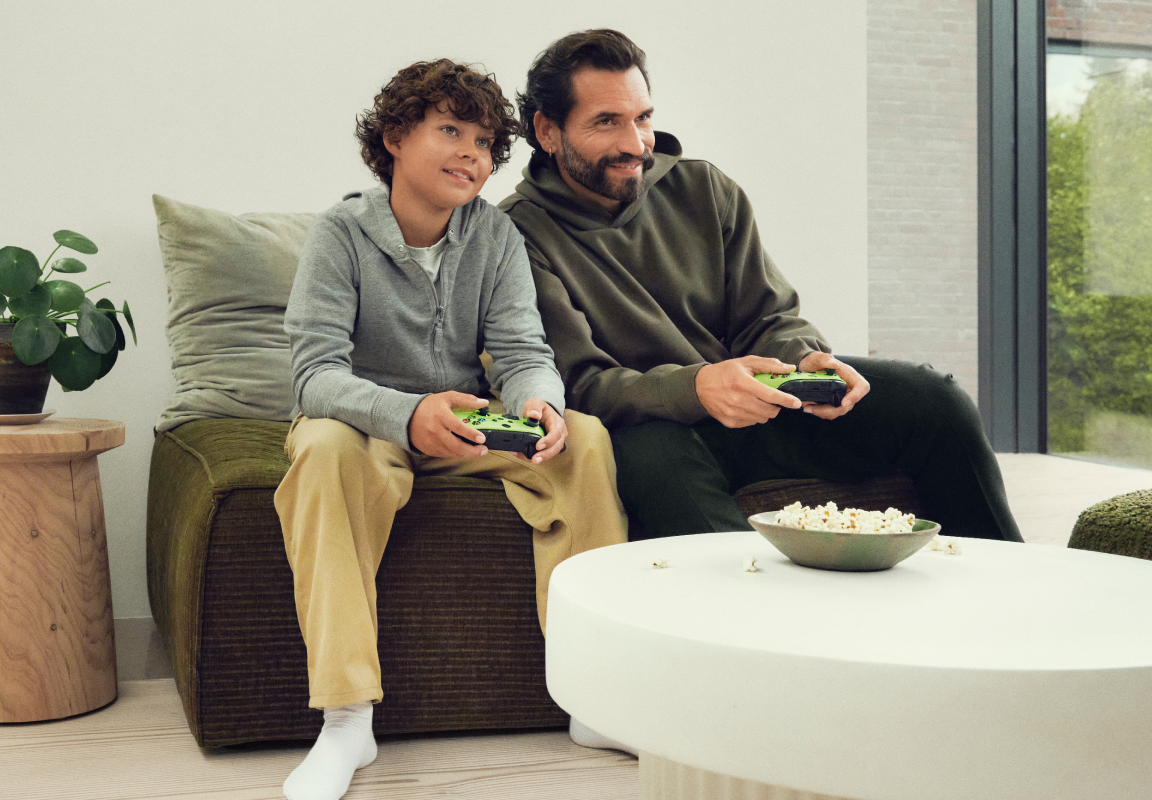 Far og søn sidder i sofaen og gamer sammen. Find dit gamingudstyr hos YouSee