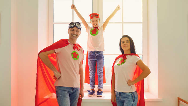 Mom and dad wear red capes with their son while standing in front of a large window in their home dressed as super heroes
