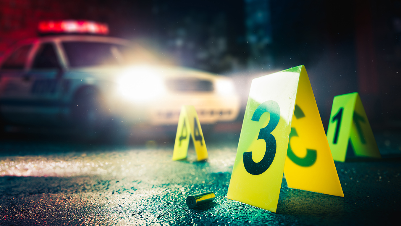 Yellow signs mark evidence at a crime scene on a street with a police car in the background with headlights on