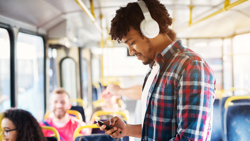 Man stands on a bus deciding which podcast to listen to while in transit wearing a plaid blue and red shirt
