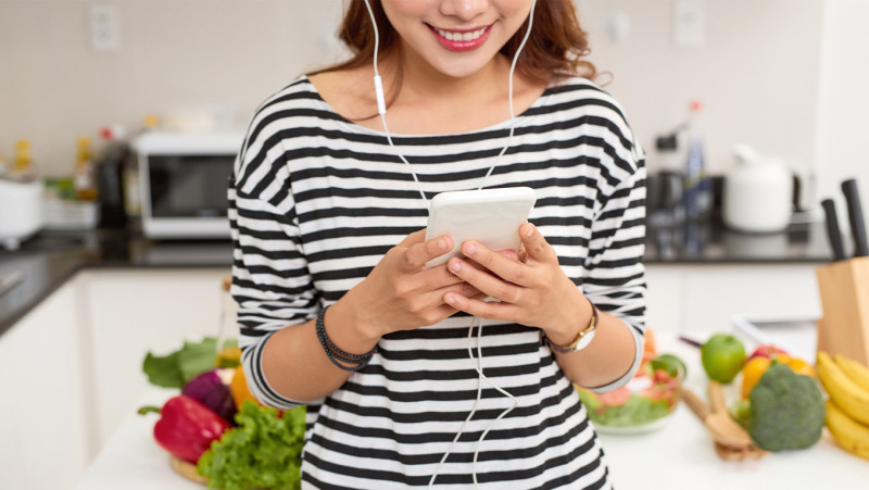 An Asian woman listens to an audiobook on her smart device with earbuds as she stands in front of a vegetable-laden cutting board