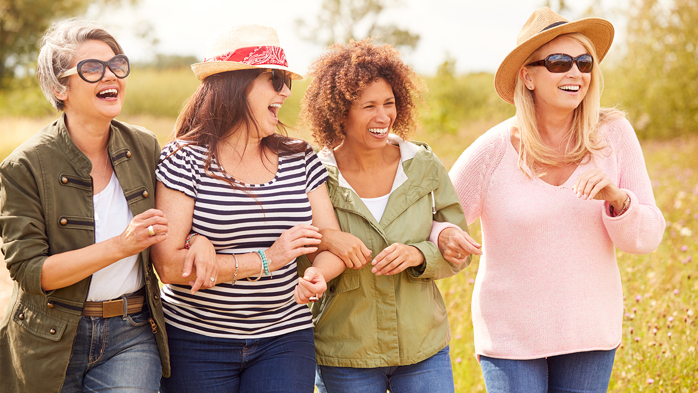 Group of mature female friends walk arm in arm through a grassy field talking and laughing