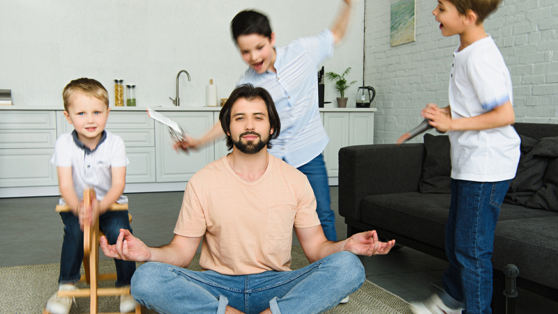 Father sits in his living room on a carpet meditating, trying to stay calm while his 3 sons run and play around him
