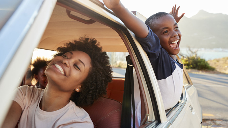 An African American family is on a road trip. The son is hanging out the back window with arms spread, smiling