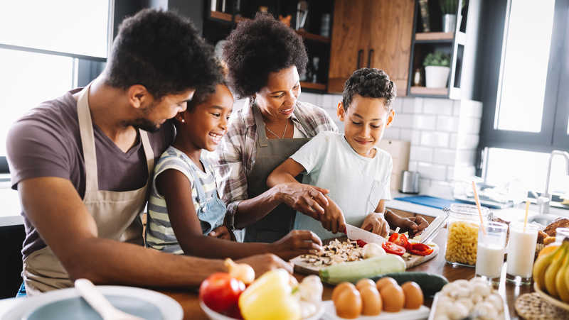 A happy family stands at a kitchen island in aprons surrounded by ingredients such as eggs and peppers, and prepares a healthy meal together