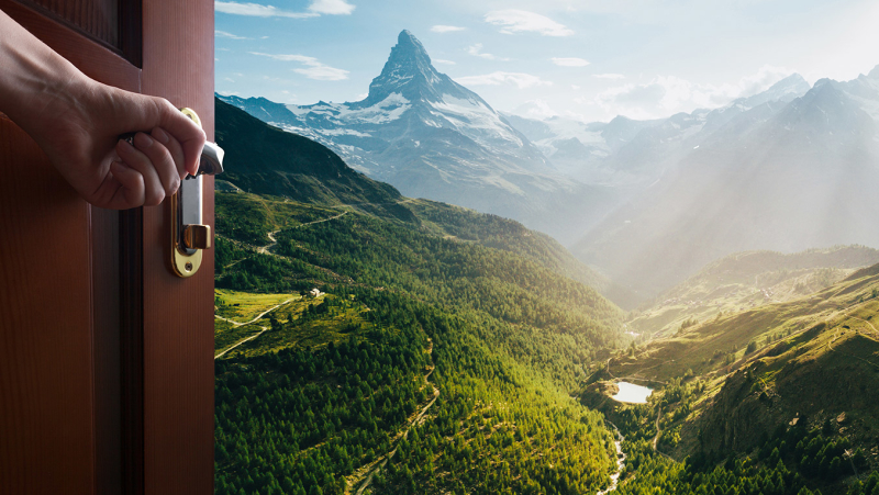 A hand opens a wooden door leading to a beautiful nature scene with green mountains and a river 