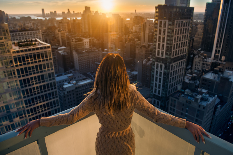 A woman stands on a balcony overlooking a city skyline at dawn