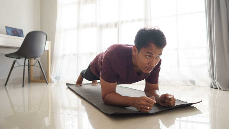 A man is holding the plank position on a yoga mat in a brightly-lit room