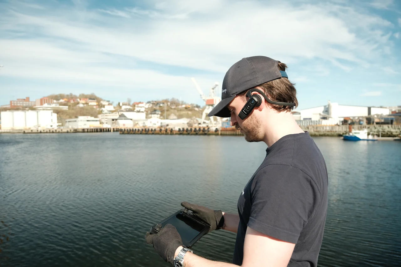 Blueye-operatør som styrer dronen fra kaia med et Bluetooth-headset, med havnen og det industrielle skylinen i bakgrunnen