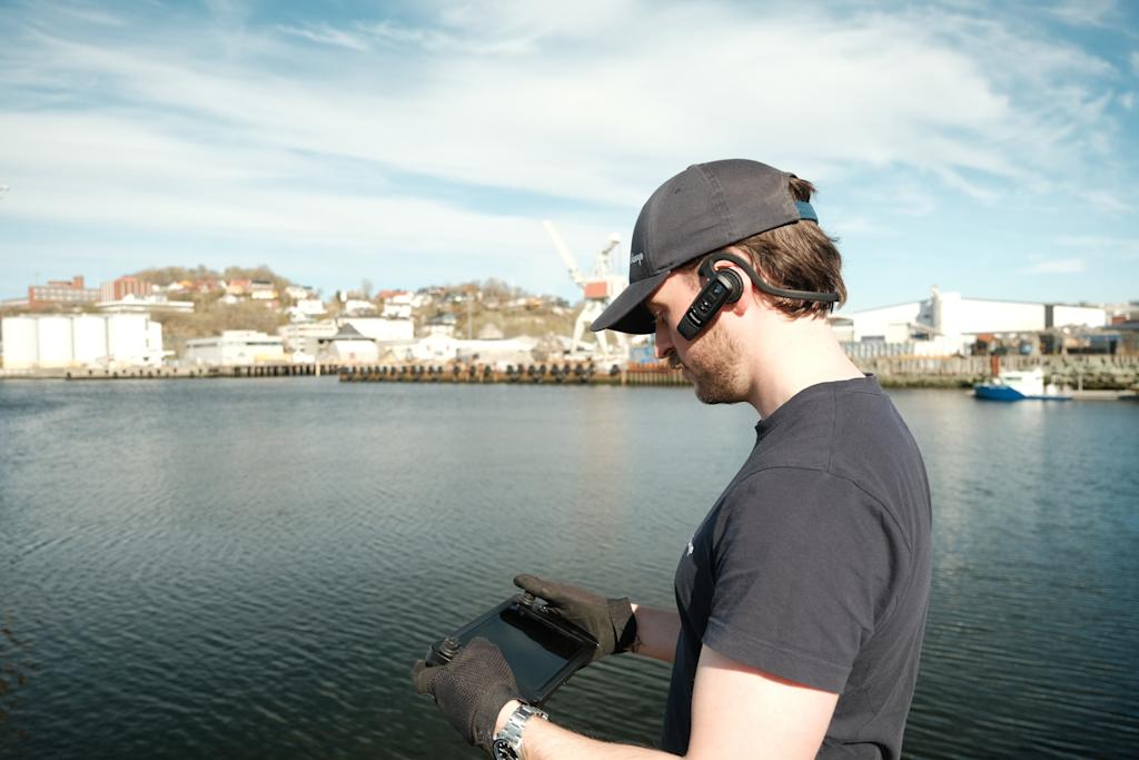 Blueye operator piloting the drone from the quay while wearing a Bluetooth headset, with the harbor and industrial skyline in the background