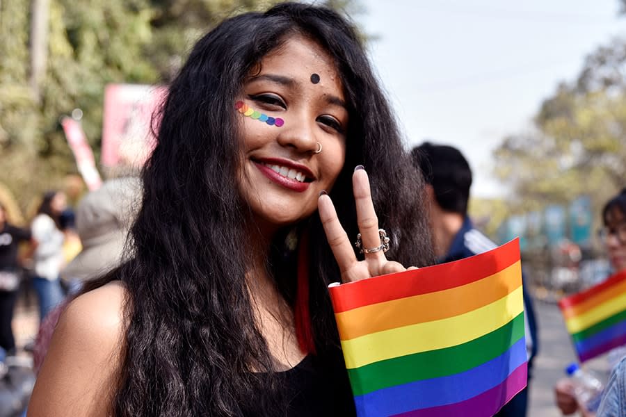 Indian girl smiling at the camera holding rainbow flag and peace sign with her fingers parade behind