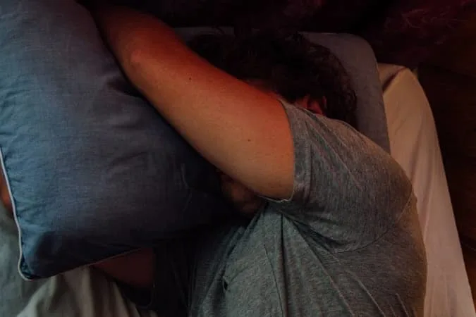 A person in a grey shirt lying in bed with their head resting on a blue pillow and their arm covering their face.