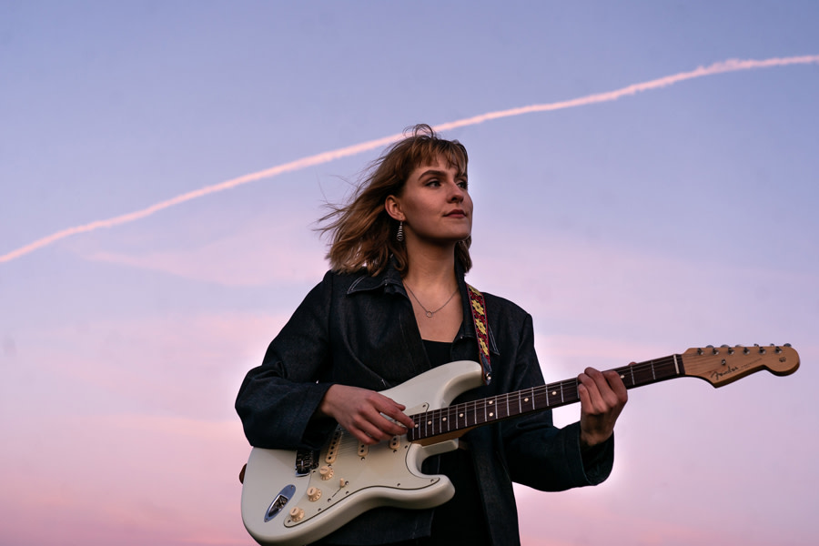 Musician Lauren Eddy at the top of a hill in front of a purple sunset holding an electric guitar