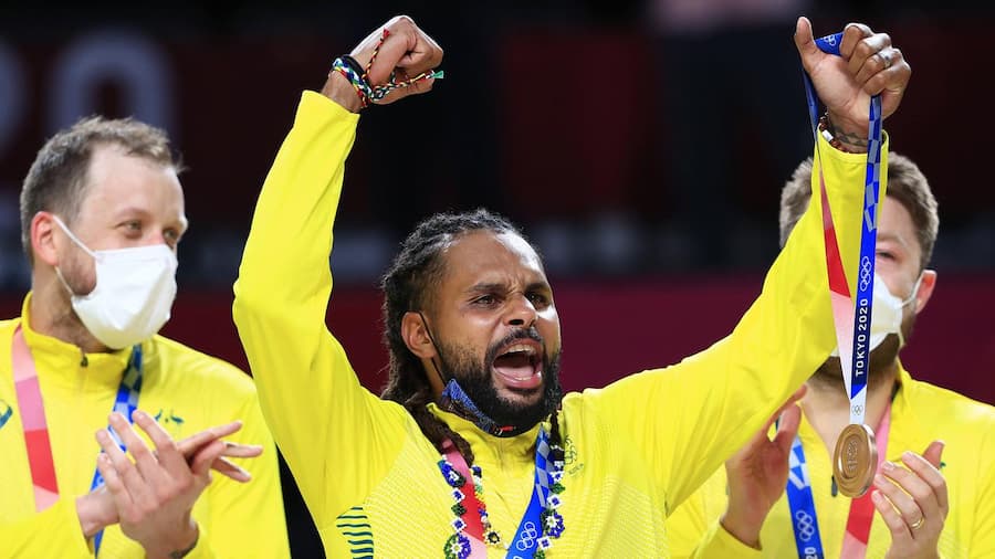 Image of Patty Mills at the Olympic Games. He has both hands in the air and is cheering. He is holding a bronze medal in one hand. 