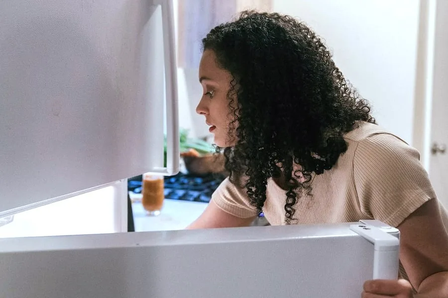 Photo of a girl leaning into an open fridge looking worried or apprehensive. The girl wears are white t-shirt and has dark shoulder length ringlets.