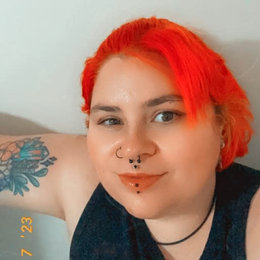 Selfie of Kai who is looking up and smiling at the camera. They have bright red hair, a tattoo on their right arm and piercings on their nose and lips.
