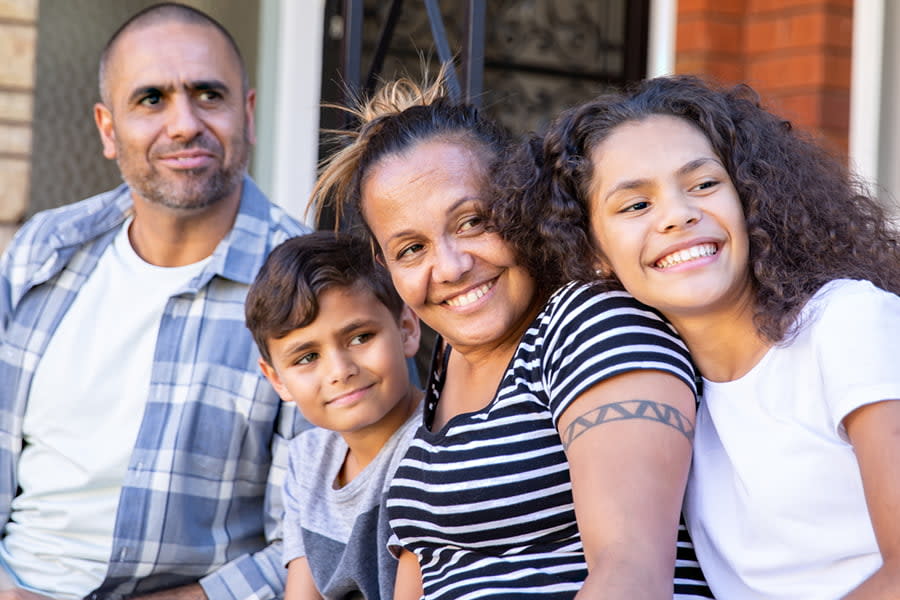 Image of an Aboriginal mother and father sitting out the front of their house with their two kids. They are all smiling and look at something off camera.